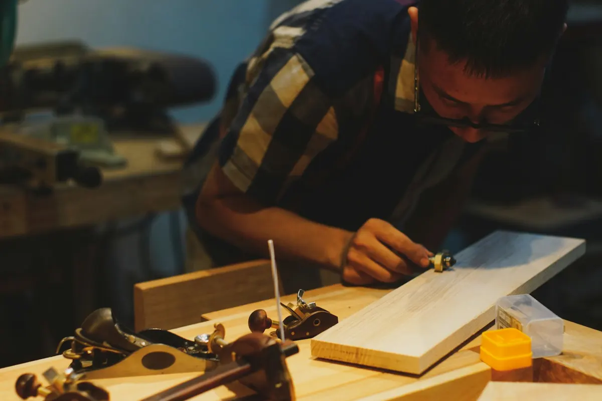 Le menuisier rabote une planche de bois clair sur l’établi, entouré de ses outils